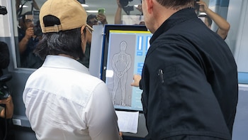 Una mujer de espaldas y un oficial examinan una pantalla táctil con la imagen de rayos X de una persona de pie y con esposas