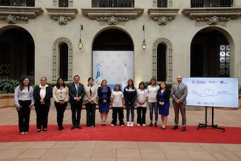 Un grupo de once personas posa sobre una alfombra roja frente a una pantalla con gráficos, dos banderas y una pared de piedra con arcos y ventanas