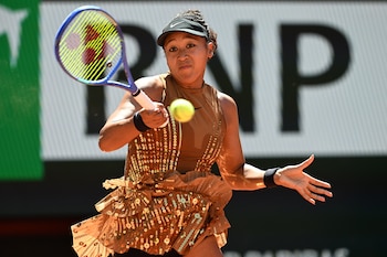 Una mujer, Naomi Osaka, golpea una pelota de tenis con una raqueta azul y amarilla en una cancha al aire libre, vistiendo un traje marrón brillante