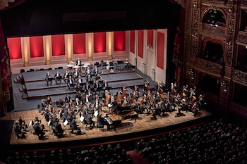 Orquesta sinfónica en un escenario de teatro con un pianista al centro, músicos con instrumentos clásicos y espectadores en la sala