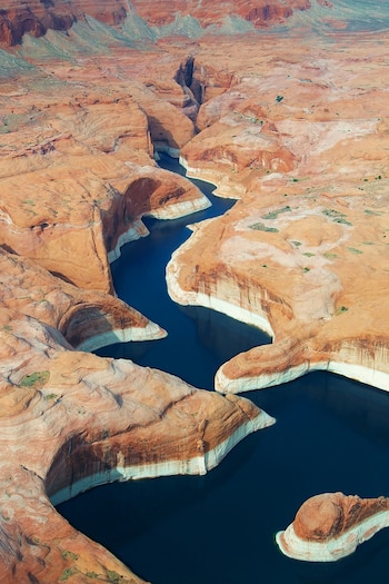Vista aérea de las aguas azules del Lago Powell serpenteando a través de cañones de arenisca roja y naranja, con evidentes líneas blancas de nivel del agua en las rocas