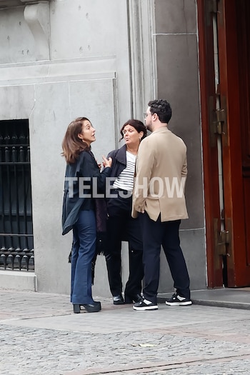 Tres personas, dos mujeres y un hombre con gafas, conversan en una acera de adoquines frente a un edificio de fachada gris y una puerta de madera marrón