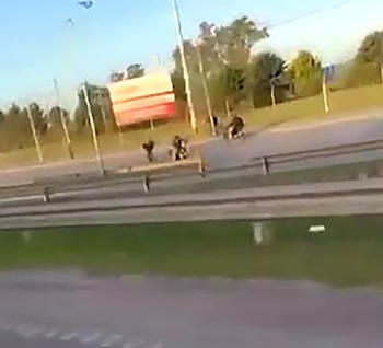 Carretera con varias motocicletas y personas a un costado de la vía, junto a carteles publicitarios y árboles