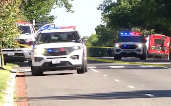 Dos vehículos SUV de policía blancos con luces de emergencia encendidas y cinta amarilla de seguridad bloquean una calle. Un autobús rojo es visible detrás. Hay árboles