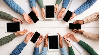 Varias manos de adultos y adolescentes rodean dispositivos móviles y tablets, algunos cuadernos y bolígrafos, sobre una mesa de madera blanca.