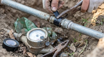 Manos con guantes manipulan una tubería de agua con cinta y alicates. Gotas de agua caen de un empalme ilegal. Un viejo contador de agua y un rollo de cinta se ven en el suelo.