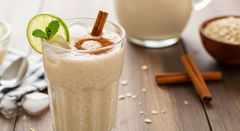 Vaso de agua de avena con hielo, decorado con una rodaja de lima, hojas de menta y canela en polvo. Al fondo, una jarra y canela en rama.