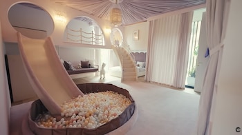 Un dormitorio con tobogán, piscina de bolas, cama, sofá, escaleras, ventana con cortinas, y techo decorado. Hay juguetes y peluches