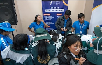 Estudiantes con uniforme y sombrero se agrupan frente a un módulo informativo atendido por tres personas con polo azul
