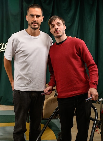 Un hombre en camiseta blanca y otro en camisa roja posan de pie. El segundo sostiene manillares de bicicleta estática. Fondo de cortina verde