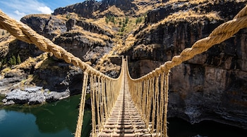 Puente colgante de cuerda y madera sobre un río. A los lados se ven montañas rocosas, vegetación seca y un cielo azul con nubes.