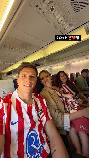 Tres adultos y un bebé sonríen en el interior de un avión. Un hombre en primer plano y dos mujeres, una con un bebé, visten camisetas del Atlético de Madrid