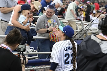 Jugadora de béisbol con uniforme a rayas y gorra azul se acerca a gradas. Una mujer sostiene a un niño con gorra. Público en un estadio, con personas filmando