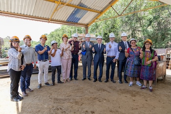 Once personas, hombres y mujeres, de pie bajo un techo metálico, todos con cascos de seguridad naranjas o blancos y haciendo un gesto de corazón con las manos