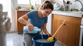 Una mujer arrodillada vierte un líquido azul de una botella de suavizante en un cubo azul con agua y una fregona.