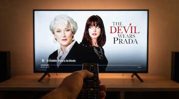 Una mano con un control remoto apunta a un televisor que reproduce el póster de 'El Diablo Viste a la Moda' con Meryl Streep y Anne Hathaway.