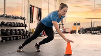 Mujer caucásica en ropa deportiva inclinada sobre un cono naranja, en un gimnasio con estantes de mancuernas, bandas de resistencia y ventanas al atardecer.