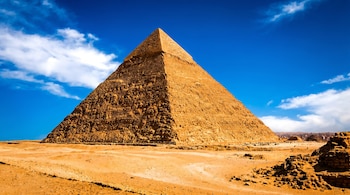 La Pirámide de Keops en Giza, Egipto, bajo un cielo azul vibrante con nubes. La estructura de piedra arenisca se alza sobre el desierto de arena amarilla.