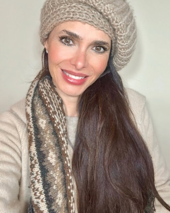Retrato de una mujer con cabello largo y oscuro, sonriendo, vistiendo un gorro de lana beige y una bufanda estampada en tonos tierra