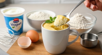 Una mano saca un trozo de bizcocho de yogur de una taza con una cuchara, con envases de yogur, harina, un huevo y un batidor en una tabla de madera.