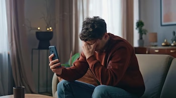 Un hombre sentado en un sofá sostiene un teléfono celular con una mano y se cubre la cara con la otra