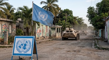 Bandera y cartel de la ONU en primer plano, convoy de vehículos blindados avanzan por una calle de tierra entre edificios bajos con grafitis y palmeras.
