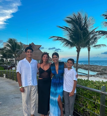 Alissa Pavano sonríe, vestida de azul, junto a dos jóvenes y un niño en un paseo marítimo, con palmeras y el mar azul de fondo bajo el cielo nublado