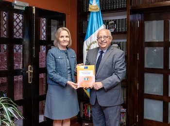Un hombre y una mujer sonríen, sosteniendo un informe naranja sobre 'Guatemala 2026'. Detrás, una bandera de Guatemala y estanterías llenas de libros