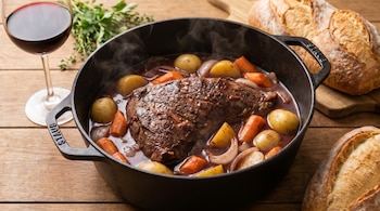 Primer plano de colita de cuadril a la cacerola humeante en una olla Staub, con patatas, zanahorias y cebolla. Se aprecian una copa de vino y pan rústico.