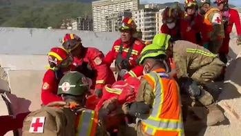 Grupo de rescatistas con uniformes rojos, cascos y chalecos de alta visibilidad asisten a una persona en una zona de escombros con edificios dañados al fondo