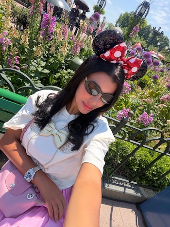 Charlotte Caniggia con orejas de Minnie, gafas de sol y camiseta blanca con lazo, posando al aire libre frente a un jardín de flores moradas y rosadas bajo el sol
