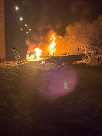 Fuego y humo intenso de un camión en llamas durante la noche. Se observa una estructura de puente, luces, árboles y pasto