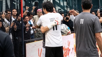 Sergio Pérez de espaldas con guantes y un balón de fútbol abraza a un hombre frente a varias personas que toman fotos con sus teléfonos.