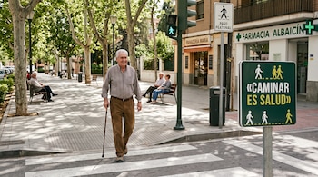Un hombre mayor con bastón cruza un paso de peatones junto a un semáforo en verde, árboles y un cartel con el texto Caminar es salud.