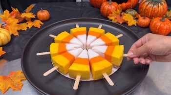 Porciones triangulares de pastel tricolor con palitos de madera en un plato negro, rodeadas de calabazas pequeñas, hojas secas y una mano.