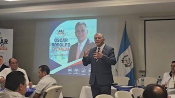 Hombre de traje habla con las manos levantadas frente a una pantalla con su fotografía y nombre, junto a una bandera de Guatemala