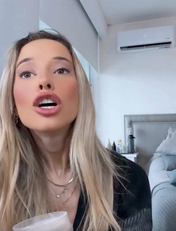 Primer plano de Zoe Bogach, mujer rubia con boca abierta, sosteniendo un vaso blanco. Fondo blanco con un aire acondicionado y parte de una cama