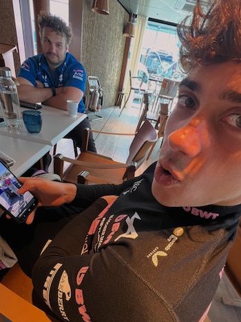 Un hombre en primer plano toma una selfie con un teléfono móvil, otro hombre con camiseta azul de pie en el fondo, una mesa y botellas
