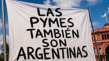 Primer plano de un cartel blanco con letras negras que dicen LAS PYMES TAMBIÉN SON ARGENTINAS. Al fondo, un edificio rosa y una bandera de Argentina ondean.