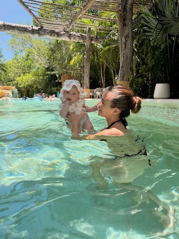 Una mujer con el pelo recogido sostiene a un bebé con un sombrero blanco de fresas en una piscina. El fondo muestra vegetación tropical y una estructura de madera