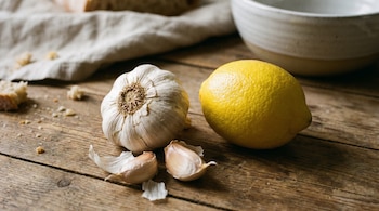 Primer plano de un bulbo de ajo entero, dos dientes de ajo y un limón amarillo sobre una mesa de madera. Al fondo, pan, un paño y un cuenco cerámico.