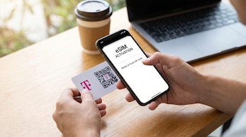Manos sostienen un smartphone con 'eSIM ACTIVATION' en pantalla y una tarjeta T-Mobile con código QR, en una mesa de madera junto a una laptop y café.