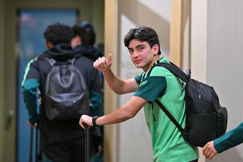 Un joven con camiseta verde de México lleva una mochila negra, toma el asa de una maleta y levanta el pulgar en un pasillo