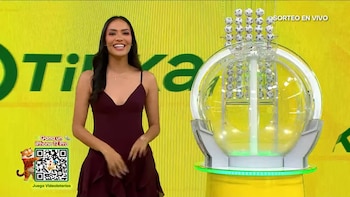 Una mujer vestida de granate permanece de pie junto a una máquina transparente de esferas sobre un fondo amarillo