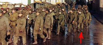 Grupo de soldados con uniformes militares de color oliva y algunos con boinas rojas marchan por una calle mojada. Al fondo se ven edificios y vehículos
