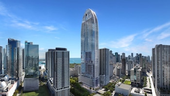 Vista aérea de la Okan Tower en Miami, un rascacielos moderno con una cima distintiva, rodeado de edificios urbanos y el océano azul al fondo