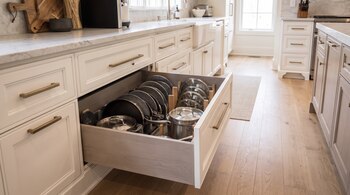 Cajón de cocina abierto que contiene sartenes en posición vertical, ollas de acero inoxidable y tapas de vidrio.