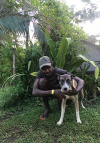 Un hombre con gorra y ropa informal posa en cuclillas junto a un perro mediano sobre césped, frente a varias plantas