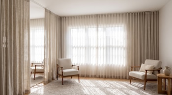 Dos sillones de madera con cojines claros, una alfombra, una mesa auxiliar con un jarrón y cortinas traslúcidas de techo a piso.