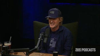 Owen Wilson lleva una gorra azul y una chaqueta oscura mientras habla frente a un micrófono en un estudio